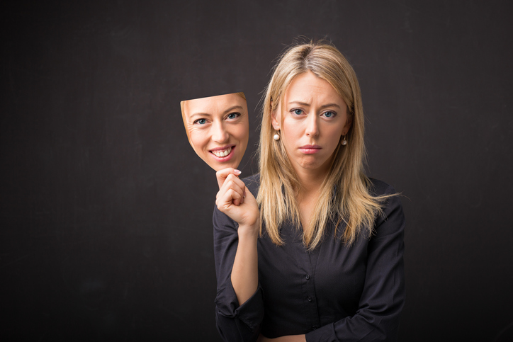 Woman holds a mask of herself smiling as she frowns.